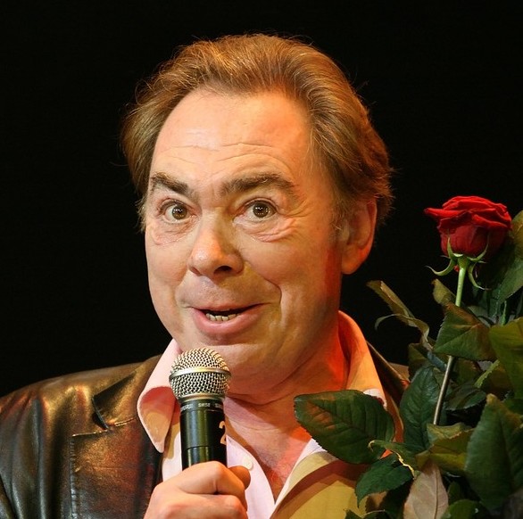 Andrew Lloyd Webber with a creepy smile speaking into a microphone while wearing a leather jacket and holding a bunch of red roses.